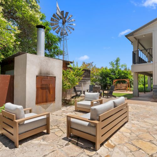 An outdoor patio with wooden lounge chairs, a stone courtyard, a tall metal windmill, and lush greenery under a blue sky.