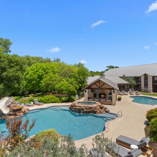 A sunny backyard resort: turquoise swimming pool, firepit patio, apartment-style building, trees, and an American flag.