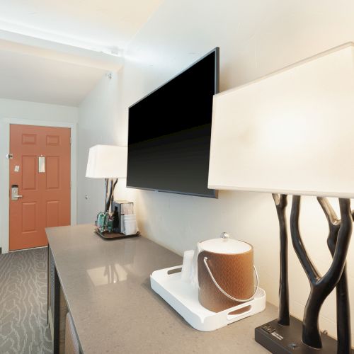 The image shows a modern hotel room with a TV, lamps, and a kettle on a wooden surface near a bed and a red door.