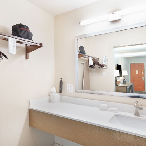 A hotel room bathroom with a sink, large mirror, and a shelf with hangers and an ironing board.