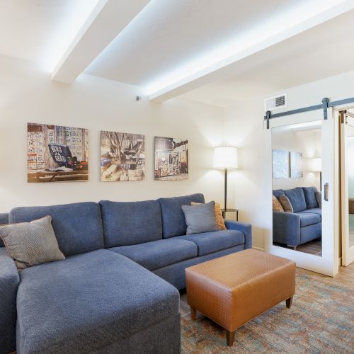 The image shows a living room with a blue sectional sofa, floor lamp, coffee table, artwork on walls, and a doorway leading to a bedroom.