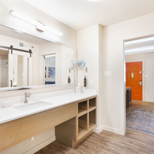 The image shows a bathroom with a long vanity, mirror, and wood flooring, next to a room with an orange door in the background.