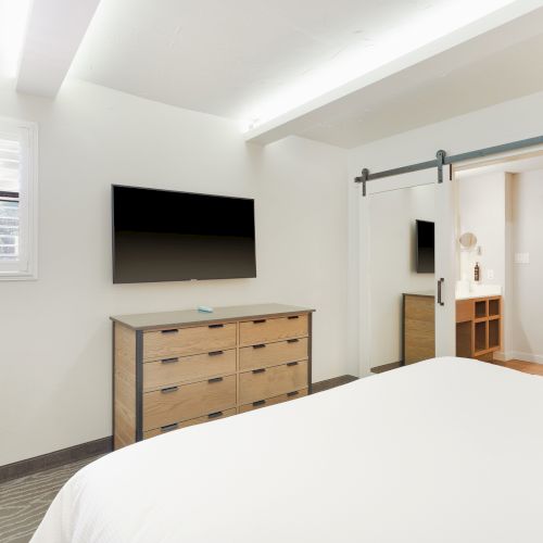 A modern bedroom with a bed, dresser, mounted TV, sliding door, and window with shutters, all in a bright, minimalistic setting.