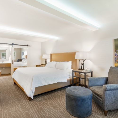 The image shows a modern hotel room with a bed, nightstands, lamps, a chair with an ottoman, a dresser, and a bathroom in the background.