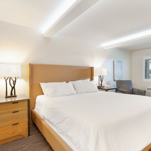 A well-lit bedroom featuring a large bed with white bedding, a wooden nightstand with a lamp, a chair, and a window with blinds.