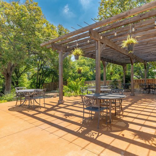 A sunny outdoor patio with a wooden pergola, metal tables and chairs, hanging plants, and lush green trees in the background, under a bright blue sky.