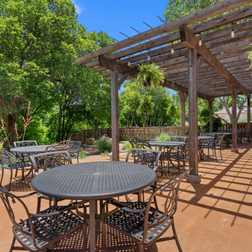 Sunny backyard patio with a wooden pergola, hanging lights, round table and chairs, and lush green trees.