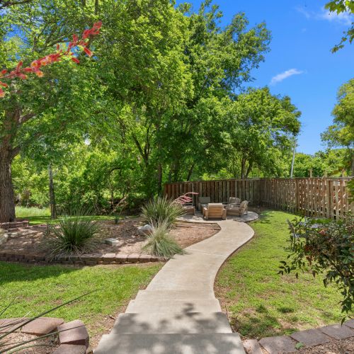 A sunny backyard garden with lush trees, blooming red flowers, a winding stone path, and patio chairs beside a wooden fence.