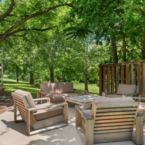 A sunny backyard patio with wooden lounge chairs and a coffee table, surrounded by lush green trees and grass.