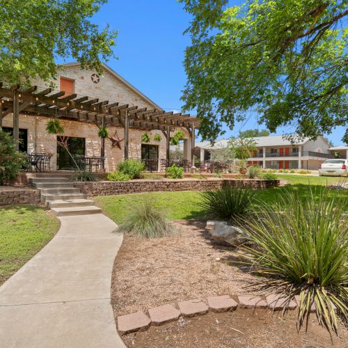 A landscaped courtyard with stone buildings, a wooden pergola with lights, outdoor seating, trees, and parked cars.