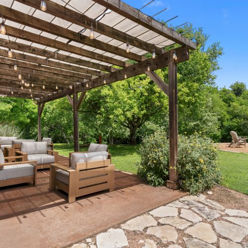 A sunny backyard patio with a wooden pergola strung with bulbs, cushioned chairs, and lush green trees.
