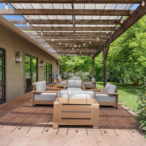 Outdoor patio with cushioned lounge chairs and a sofa under a wooden pergola with warm hanging bulbs, surrounded by lush green trees.
