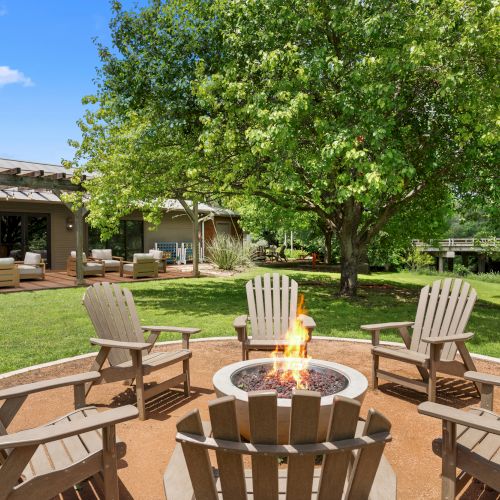 A sunny backyard patio with Adirondack chairs around a crackling fire pit, leafy trees, and a deck with outdoor seating.