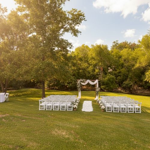 An outdoor wedding setup with arranged chairs, a decorative arch, and a table on a grassy area surrounded by trees under a sunny sky.