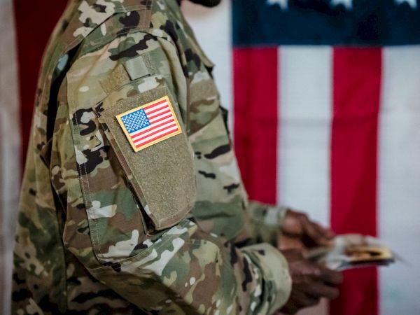 A soldier in camouflage with an American flag patch counts money against a backdrop of the U.S. flag, wartime-like scene.