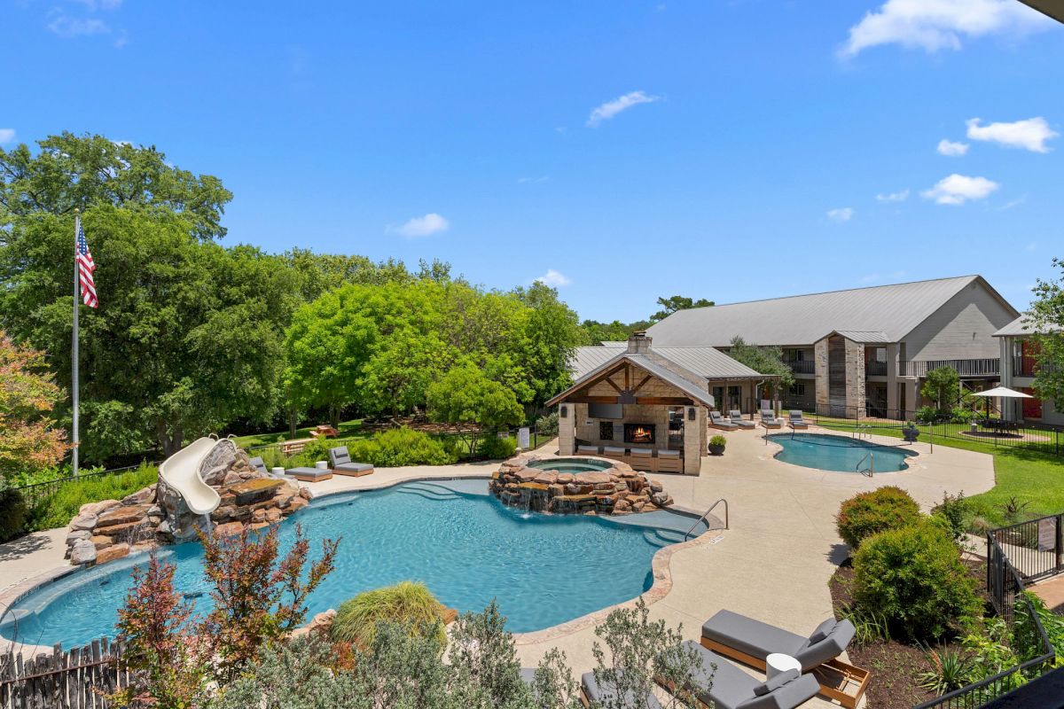 A large pool area with a water slide, pavilions, and lounge chairs surrounded by trees and a building in the background.