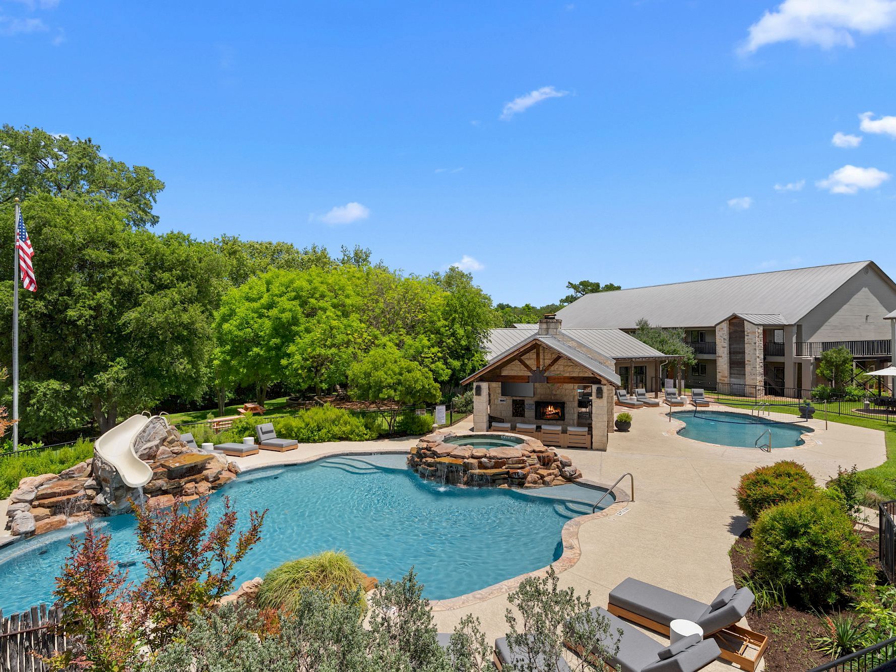 A large pool area with a water slide, fireplace, and lounge chairs surrounded by trees and a building in the background.