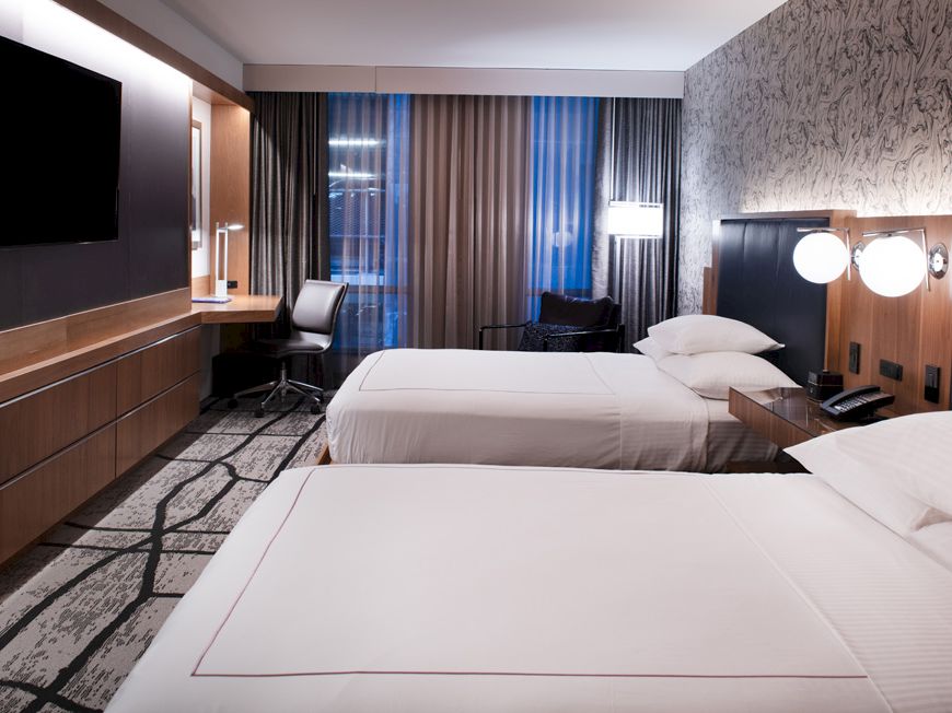A modern hotel room with two beds, sleek wood panels, floor-to-ceiling curtains, and wall-mounted TVs.
