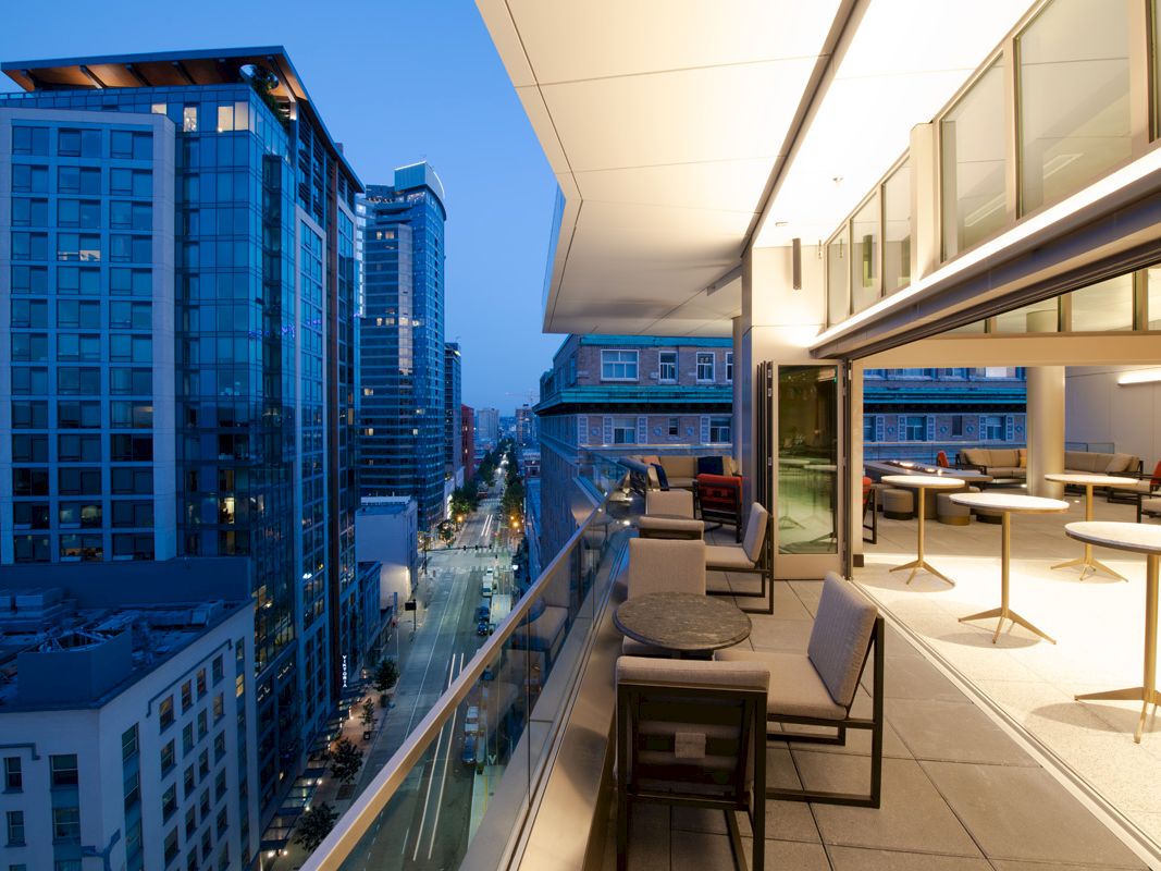 A modern high-rise balcony lounge overlooks a glowing city street between tall office buildings.